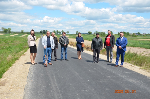 Osiem osób stojących na świeżo wyasfaltowanej drodze, w tle zielone pola i niebieskie niebo z chmurami.