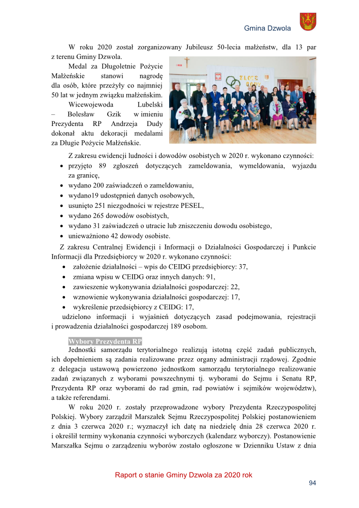 Grupa seniorów w formalnym ubiorze siedzi i stoi w sali z jasnymi ścianami, na ścianie krzyż i dekoracja z napisem "Złote Gody".