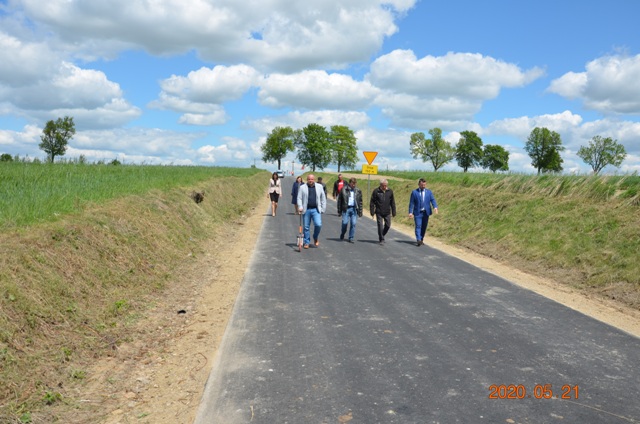 Grupa osób idąca po świeżo wyasfaltowanej drodze otoczonej zielonymi skarpami pod niebieskim niebem z białymi chmurami.