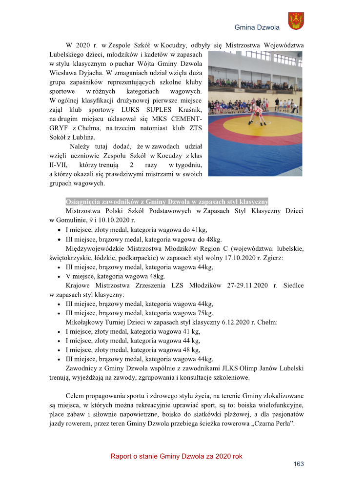 Zawodnicy na macie do zapasów w hali sportowej, wokół nich widzowie na trybunach, kolorowa mata z czerwonym i żółtym kołem.