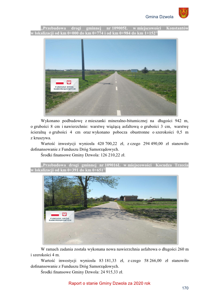 Droga asfaltowa z linią przerywaną pośrodku, po bokach zielone pola, słup energetyczny oraz tablica informacyjna; druga droga asfaltowa z drzewami i budynkami w tle, niebo bezchmurne.