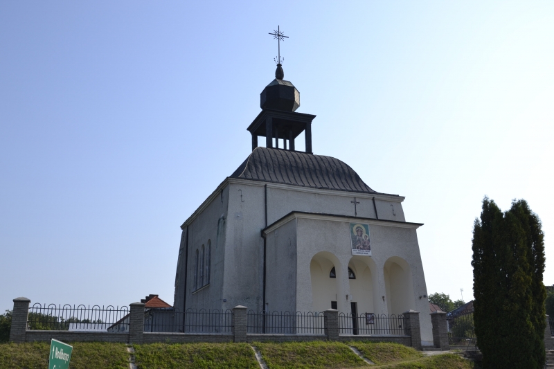Biała kaplica z ciemnym dachem i wieżyczką zakończoną krzyżem, otoczona niskim murem z metalowym ogrodzeniem, na tle jasnego nieba i zieleni.