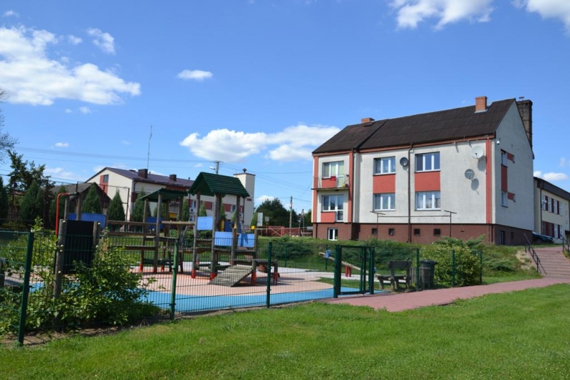 Plac zabaw z kolorowymi urządzeniami, ogrodzony zielonym płotem, obok trawiasty teren i dwupiętrowy budynek mieszkalny pod niebieskim niebem.