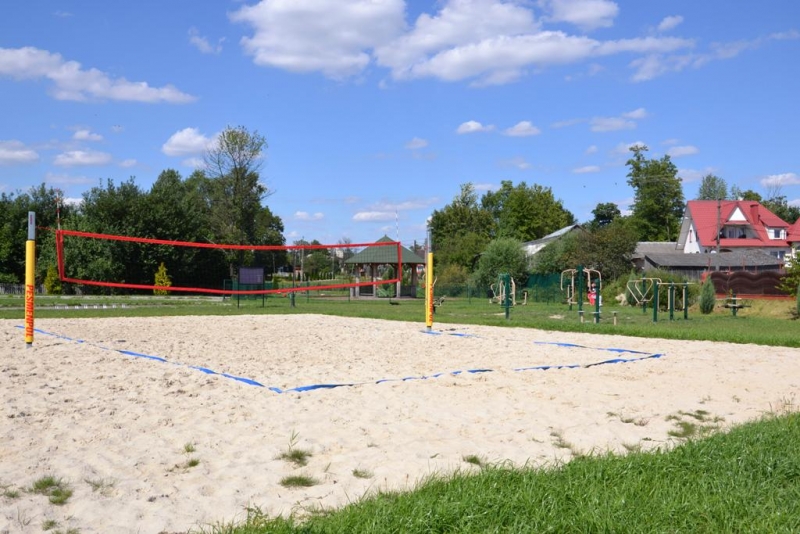 Boisko do siatkówki plażowej z czerwono-żółtą siatką na piasku, w tle zielone drzewa, trawa i zabawki na placu zabaw, pod błękitnym niebem z białymi chmurami.