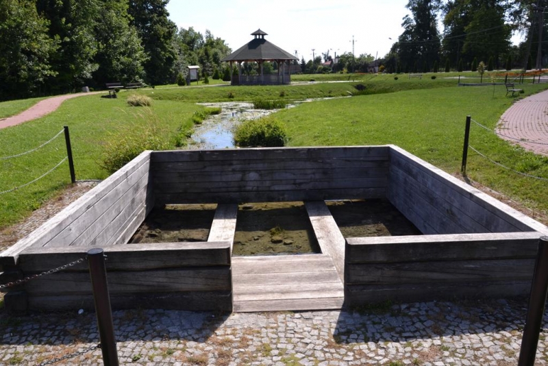 Kwadratowa, drewniana, otwarta konstrukcja na kamiennej nawierzchni, w tle zielona trawa, ścieżki, drzewa i altana z ciemnym dachem.