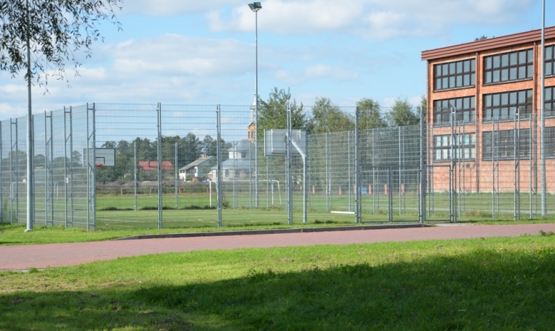 Boisko otoczone metalowym ogrodzeniem, dwa słupki do koszykówki, zielona trawa, budynek z dużymi oknami, niebieskie niebo z chmurami.