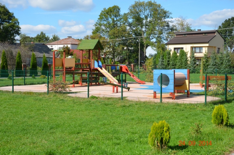 Plac zabaw z drewnianą konstrukcją, dwoma zjeżdżalniami i tunelem, ogrodzony zielonym metalowym płotem, na tle zielonej trawy, drzew i domów.