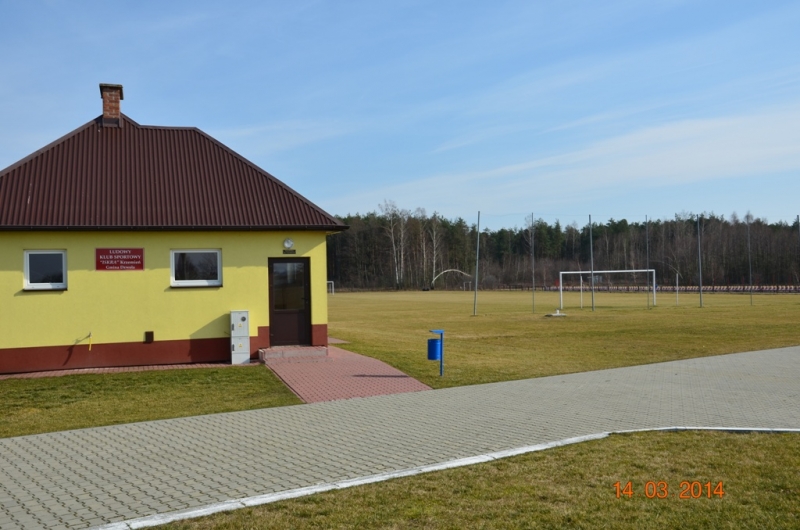 Żółty budynek z brązowym dachem obok chodnika, za nim zielone boisko piłkarskie z bramką, w tle las i błękitne niebo.