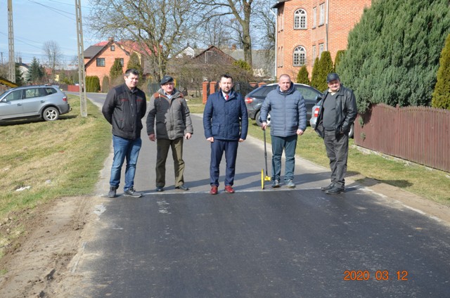 Pięciu mężczyzn stoi na nowo wyasfaltowanej drodze w słoneczny dzień, w tle domy, samochody i drzewa.