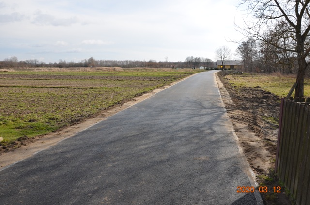 Asfaltowa droga biegnąca przez pola z resztkami zielonych roślin, po prawej stronie drzewa bez liści i drewniane ogrodzenie.