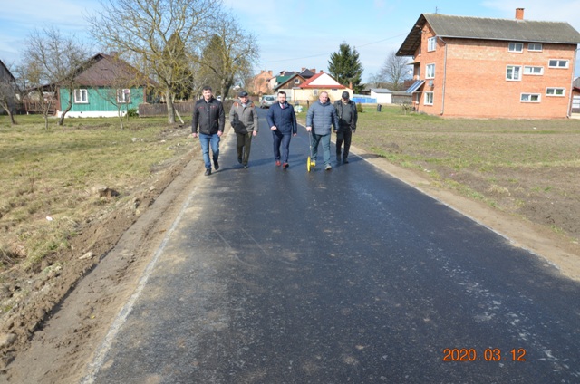 Pięć osób idących po świeżo wyasfaltowanej drodze, po bokach trawniki i domy, niebieskie niebo z lekkimi chmurami.