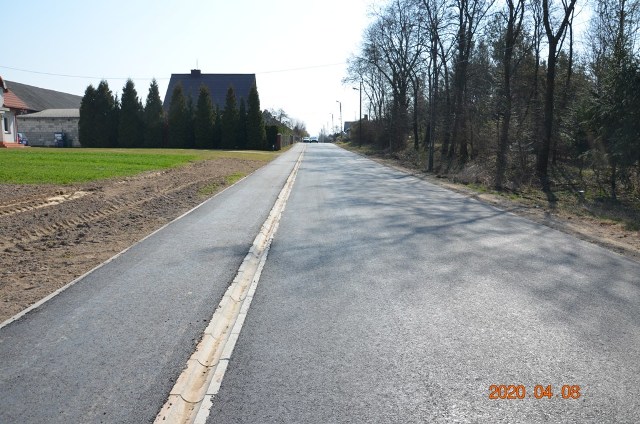 Asfaltowa droga z chodnikiem, po lewej trawa i domy, po prawej drzewa bez liści, widok w słoneczny dzień, data 2020 04 06 w prawym dolnym rogu.