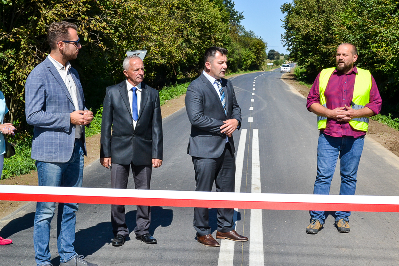Czterech mężczyzn stoi na nowo wyremontowanej drodze, przed białą i czerwoną taśmą, w tle zielone drzewa i biały samochód.