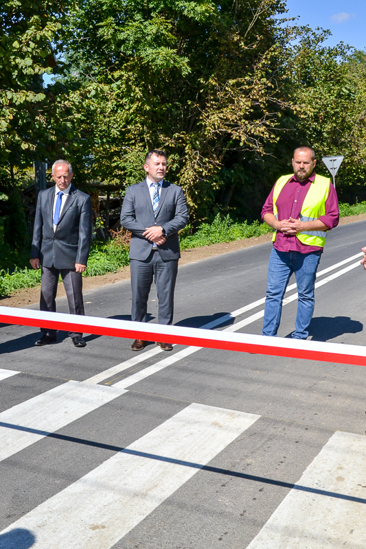 Trzech mężczyzn stojących na drodze z pasami, przed biało-czerwoną taśmą, w tle zielone drzewa i znak drogowy.