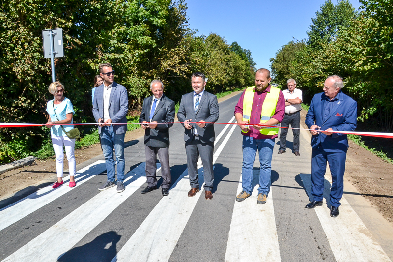 Sześć osób stoi na pasach na drodze, trzymając biało-czerwoną wstęgę, jedna z nich w kamizelce odblaskowej, w tle drzewa i niebieskie niebo.