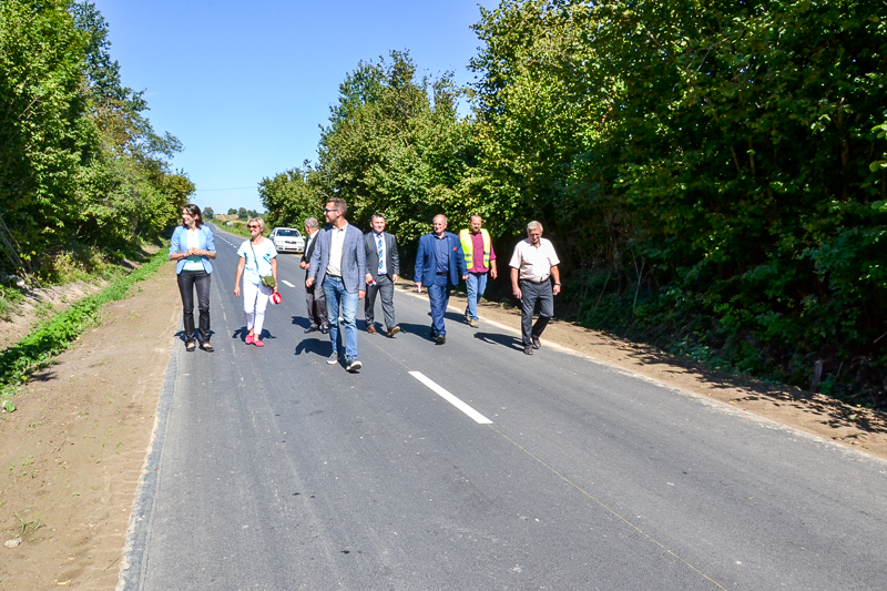 Grupa ludzi idących po nowej, asfaltowej drodze, po bokach zielone drzewa i krzewy, na niebie bezchmurne, jasnoniebieskie tło.
