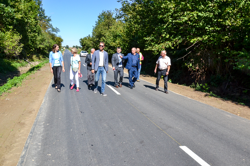 Grupa ośmiu osób idzie nowo wyremontowaną, dwupasmową drogą otoczoną zielonymi drzewami pod jasnym, niebieskim niebem.
