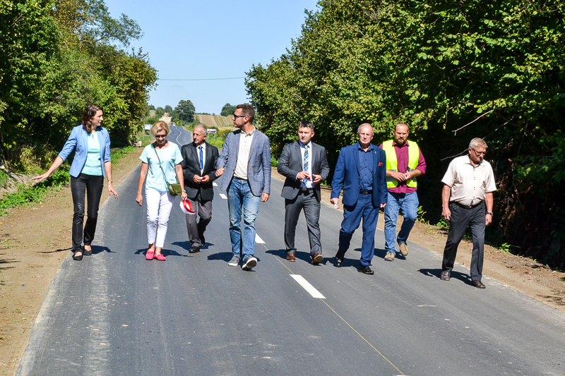 Osiem osób idących po nowo wyasfaltowanej drodze, otoczonej zielonymi drzewami i krzewami, pod jasnym, błękitnym niebem.