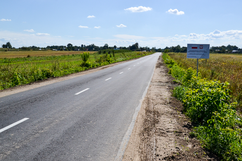Asfaltowa droga z białymi liniami na środku, po bokach pola uprawne i zielone krzewy, niebieskie niebo z kilkoma chmurami, po prawej tablica informacyjna.
