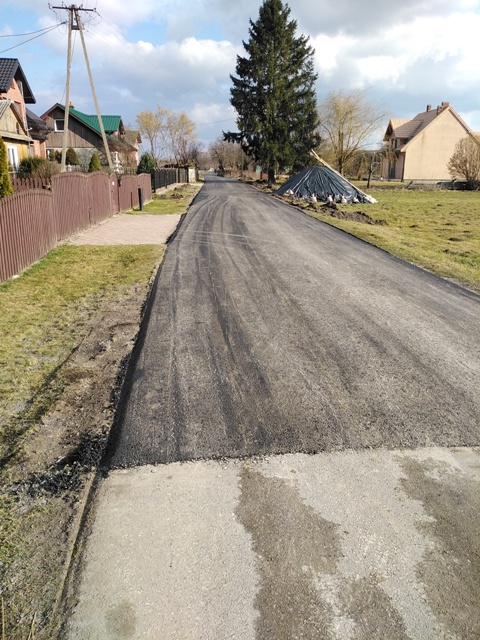Ulica o nowej, ciemnej nawierzchni asfaltowej między domami, po lewej drewniany płot, po prawej trawa i sterta przykryta czarną folią, na tle drzew i nieba z chmurami.