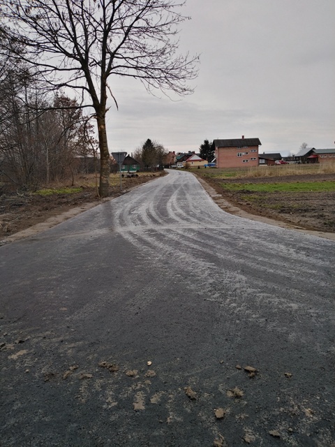 Wąska, świeżo utwardzona droga wsiowa z widocznymi śladami opon, po lewej stronie nagie drzewa, po prawej polana i budynki mieszkalne pod szarym niebem.