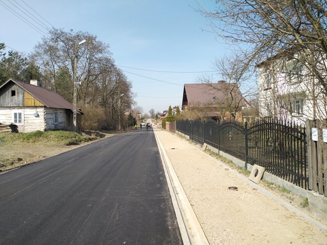 Ulica z nowym, czarnym asfaltem, po prawej stronie żwirowy chodnik, metalowe ogrodzenie i domy, po lewej stronie stare, drewniane budynki i drzewa bez liści.