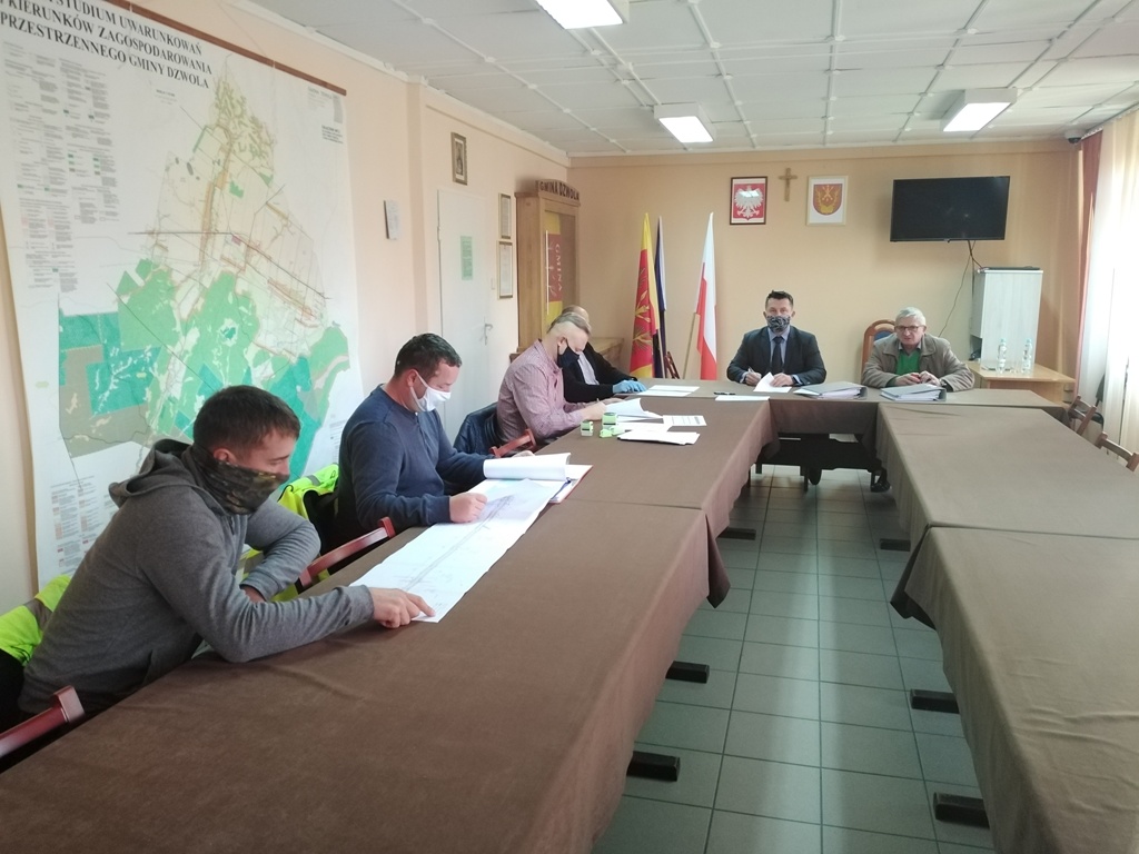 Pięć osób w maseczkach siedzi przy stołach z brązowymi obrusami, z dokumentami, na ścianie mapa, flagi Polski i herb, telewizor i krzyż.