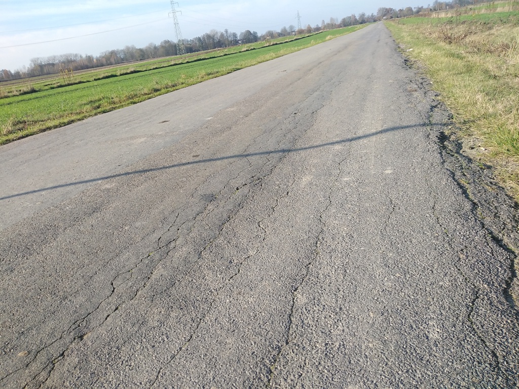 Popękany asfaltowy wiejski trakt wśród zielonych pól pod jasnym niebem, z widocznymi słupami energetycznymi w tle.