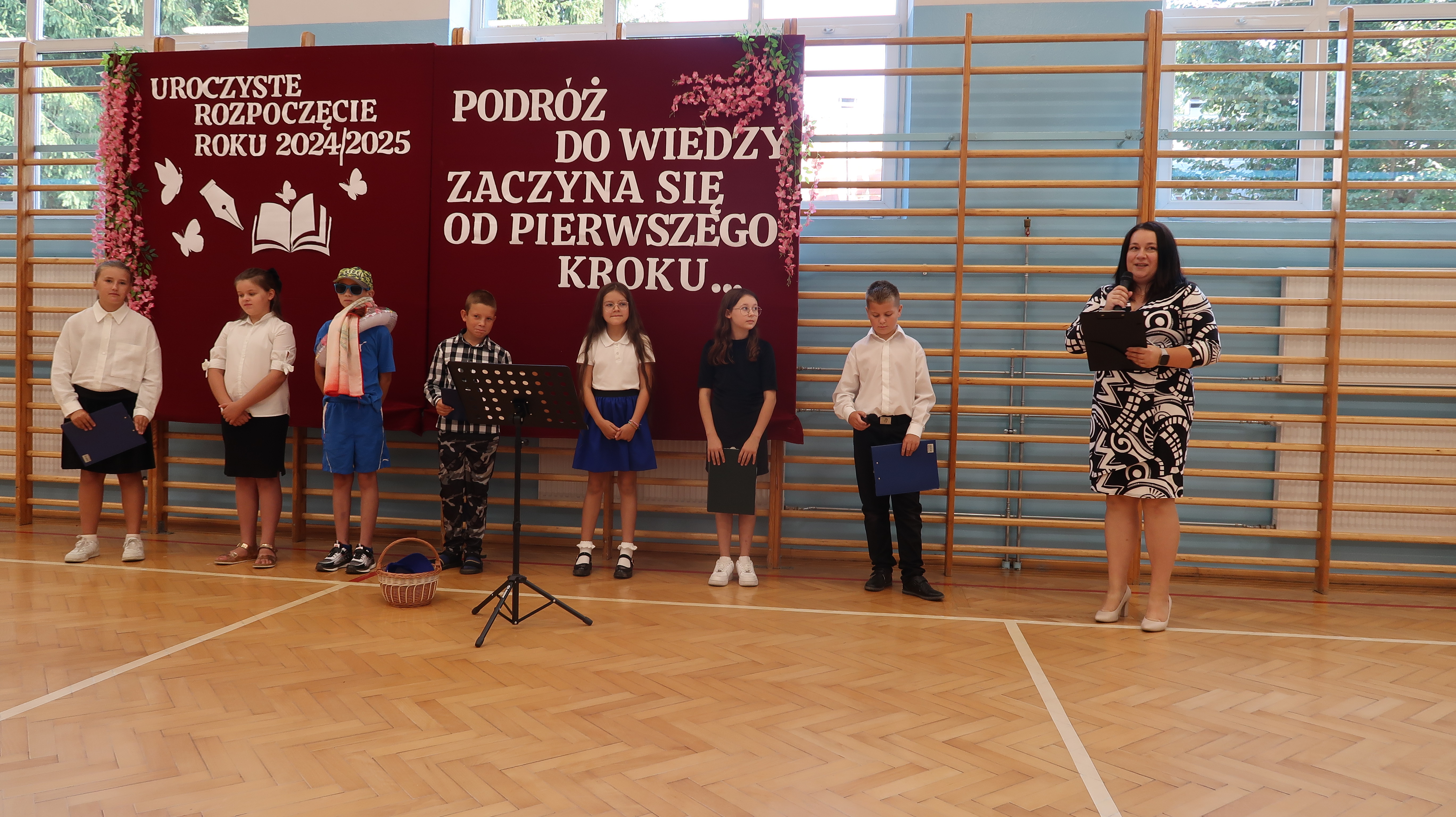 Siedmioro dzieci i kobieta z mikrofonem stoją na parkiecie hali sportowej przed bordowym banerem z białym napisem i różowymi kwiatami, za nimi drewniane drabinki.