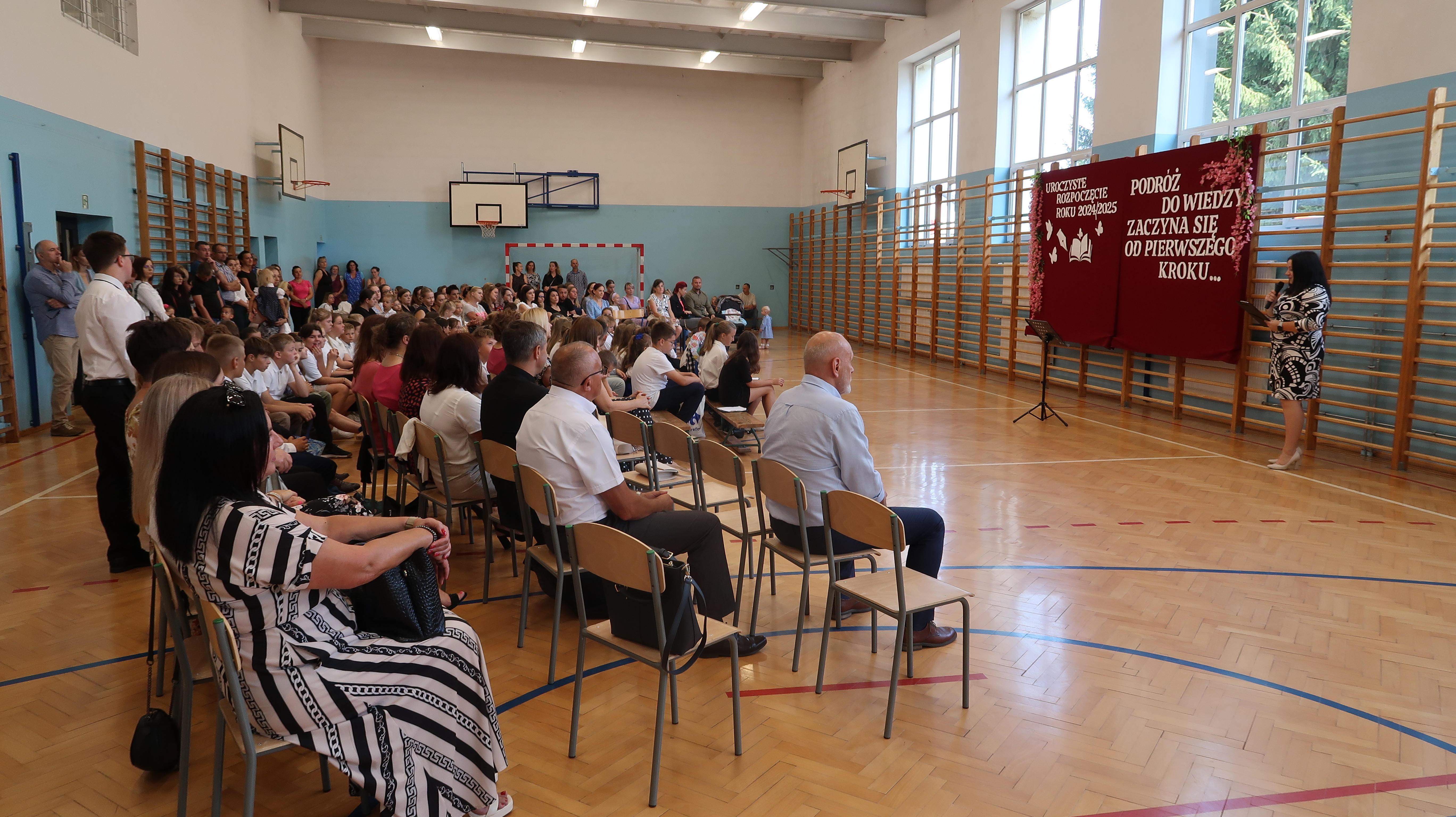 Sala gimnastyczna z drewnianą podłogą, niebiesko-białe ściany, dzieci i dorośli siedzą na krzesłach, kobieta przy tablicy z czerwonym tłem i białym napisem.