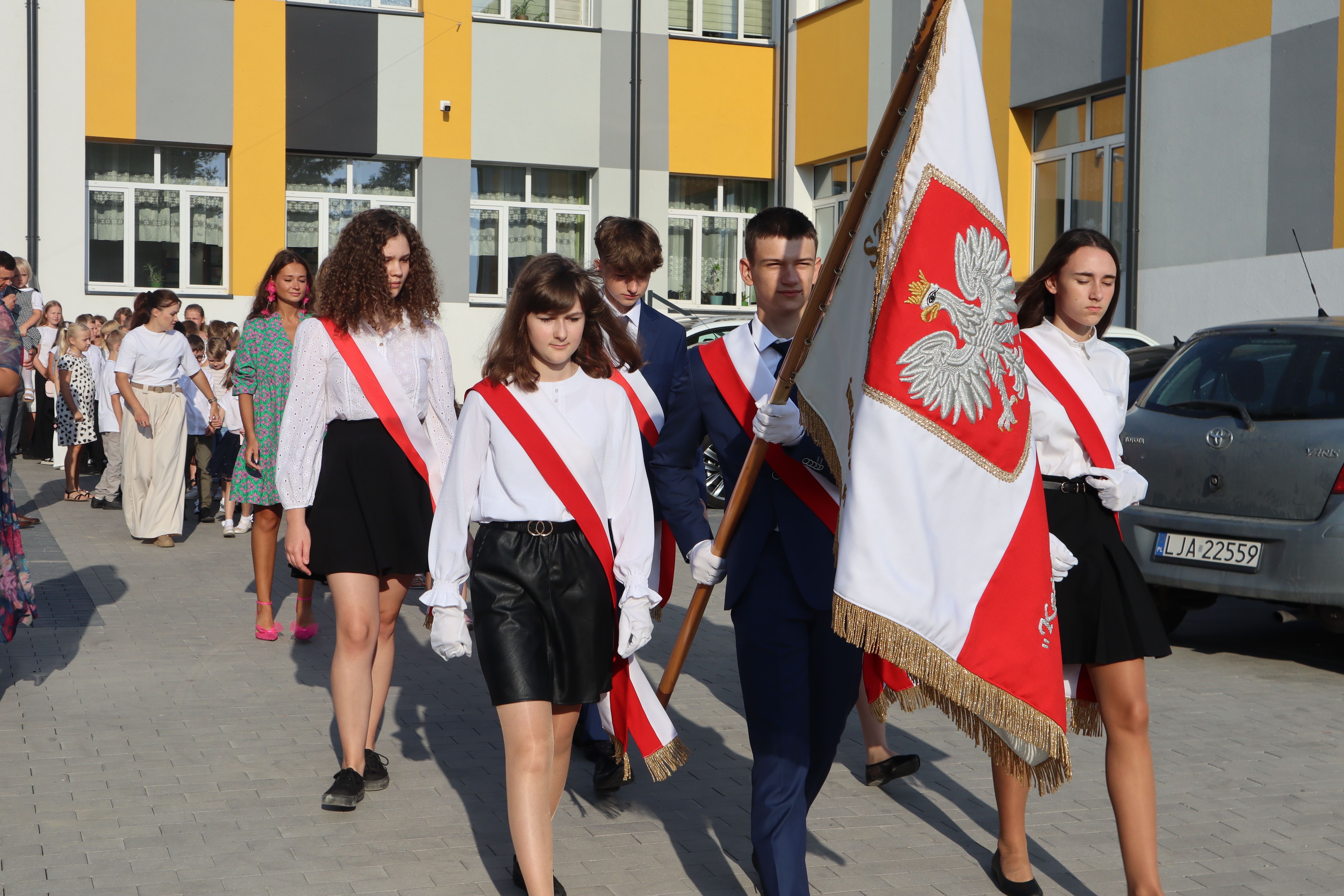 Grupa nastoletnich uczniów ubranych w białe koszule, czerwone szarfy i czarne spódnice lub spodnie, idących w pochodzie na tle żółto-szarego budynku szkoły.