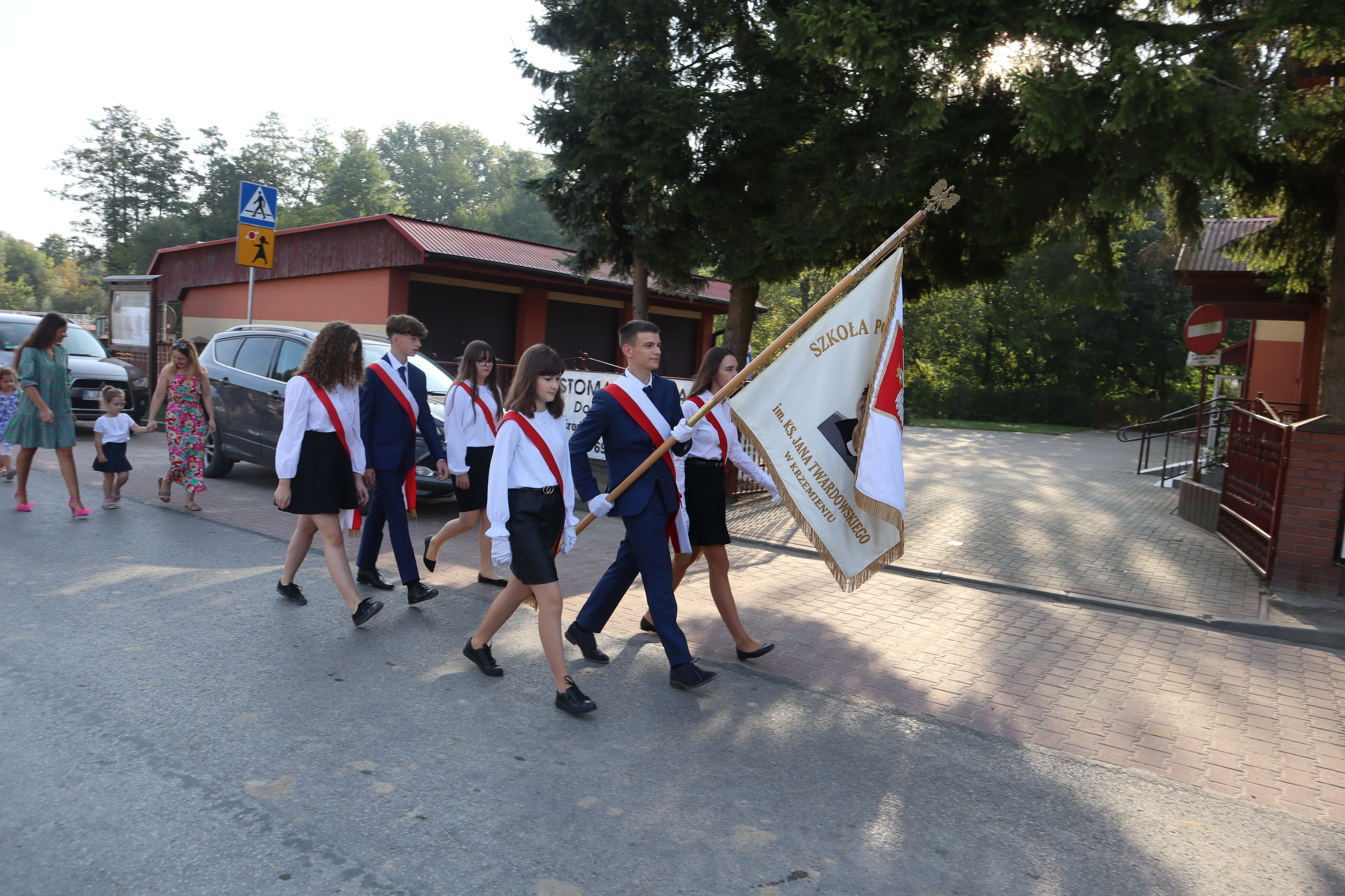 Grupa młodzieży w mundurkach szkolnych z czerwonymi szarfami, chłopak niesie biało-czerwoną flagę z napisem, idą ulicą, w tle samochody, drzewa i budynki.
