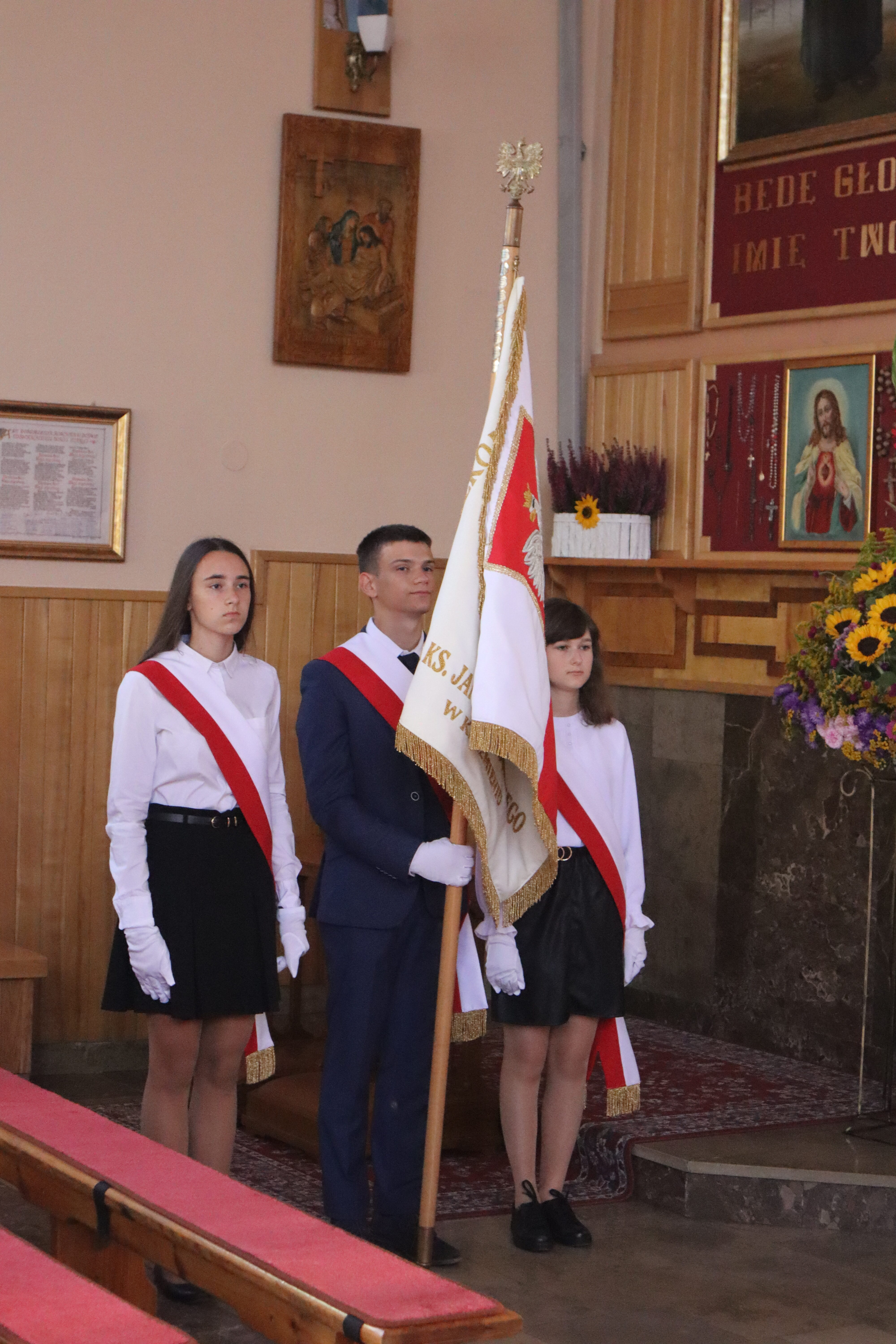 Trzy młode osoby w białych koszulach, czerwonych szarfach i białych rękawiczkach, chłopak w garniturze trzymający biało-czerwoną flagę ze złotym orłem.