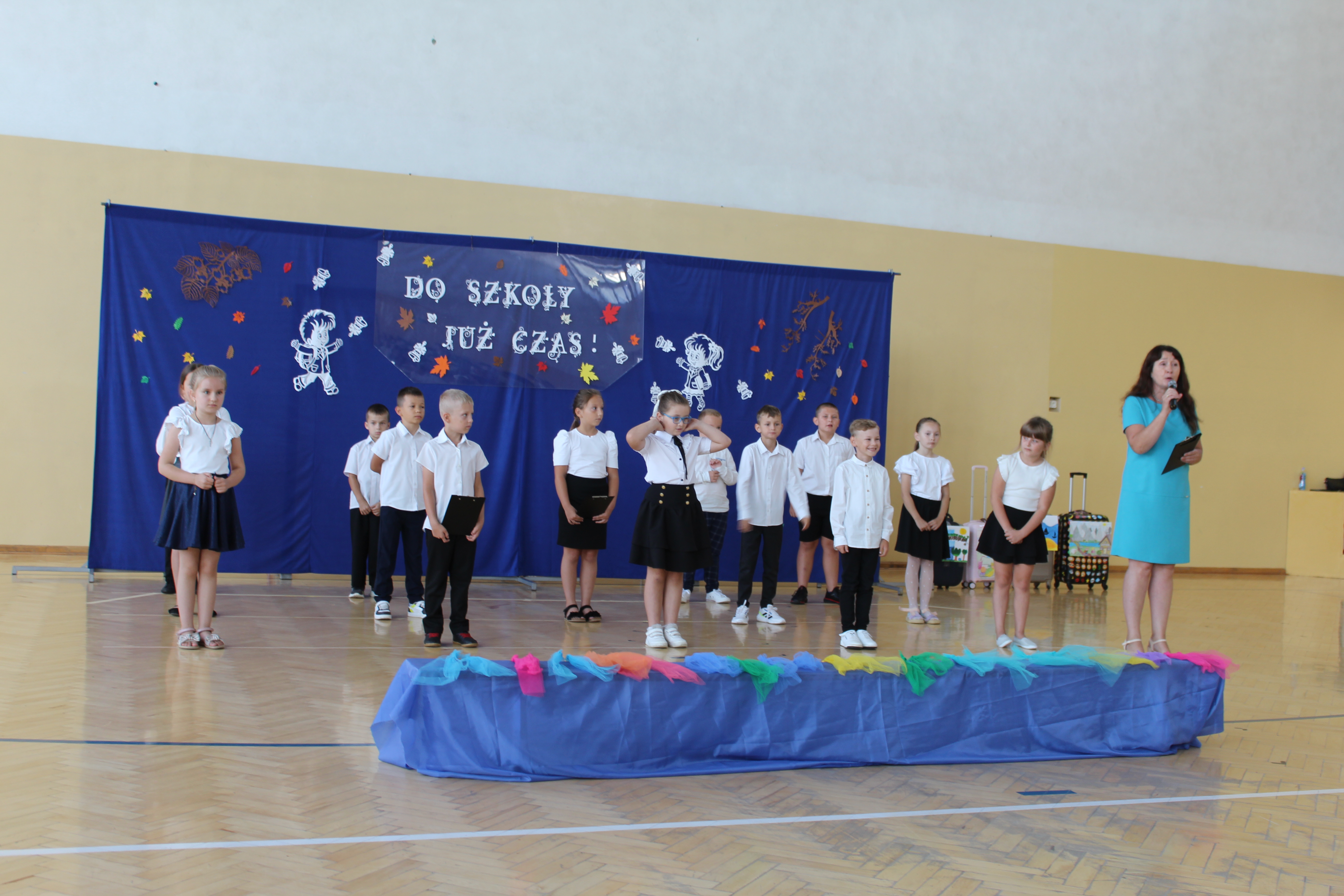 Grupa dzieci w białych koszulach i ciemnych spodniach lub spódnicach na sali gimnastycznej, z niebieskim tłem i napisem "Do szkoły już czas!", obok kobieta w niebieskiej sukience z mikrofonem.