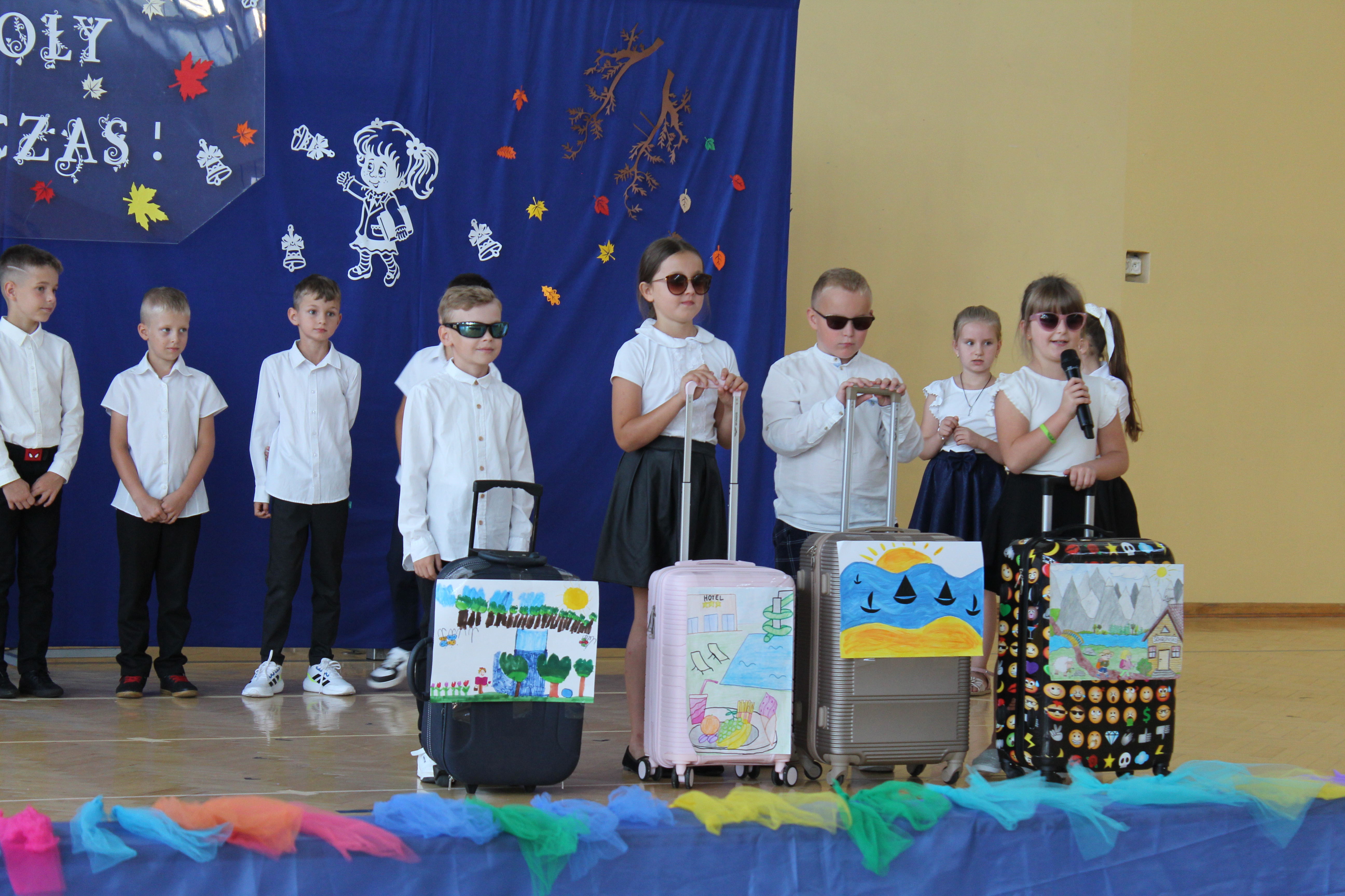 Grupa dzieci w białych koszulach i ciemnych spodniach lub spódnicach na scenie z kolorowymi walizkami ozdobionymi rysunkami, na niebieskim tle dekoracje z jesiennymi liśćmi.