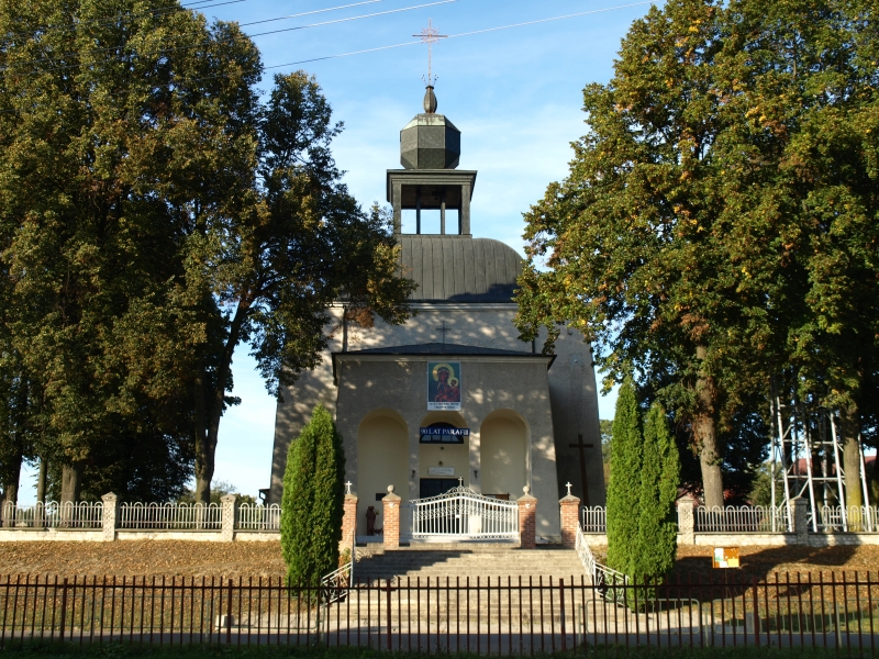 Szara murowana kaplica z wieżą, otoczona wysokimi drzewami, z białą bramą i schodami prowadzącymi do wejścia.