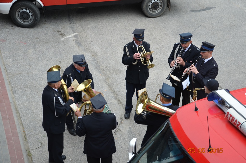 Osiem osób w mundurach grających na instrumentach dętych blaszanych i drewnianych, stojących na szarej nawierzchni obok czerwonego pojazdu strażackiego.