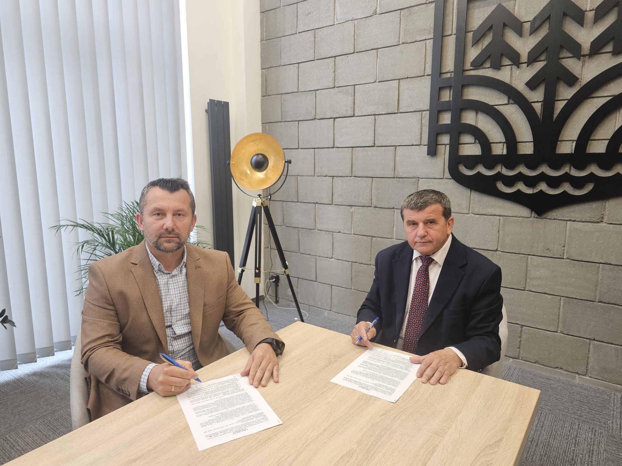 Dwaj mężczyźni w marynarkach siedzą przy drewnianym stole, podpisując dokumenty, za nimi szara ściana z czarną dekoracją i stojąca lampa na trójnogu.