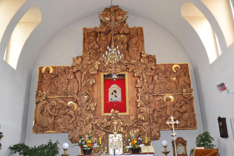 Ołtarz z drewnianymi rzeźbami przedstawiającymi sceny religijne, złote elementy dekoracyjne, czerwony obrus w centralnej części, białe ściany i okna łukowe.