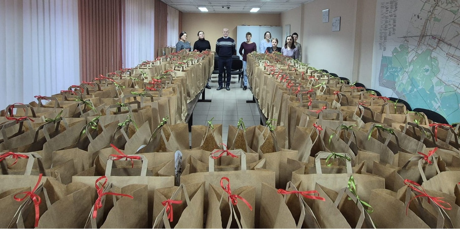 Wąski pokój z dużą ilością brązowych papierowych torebek z czerwonymi i zielonymi kokardkami na stołach, z tyłu stoi osiem osób, po prawej mapa na ścianie.