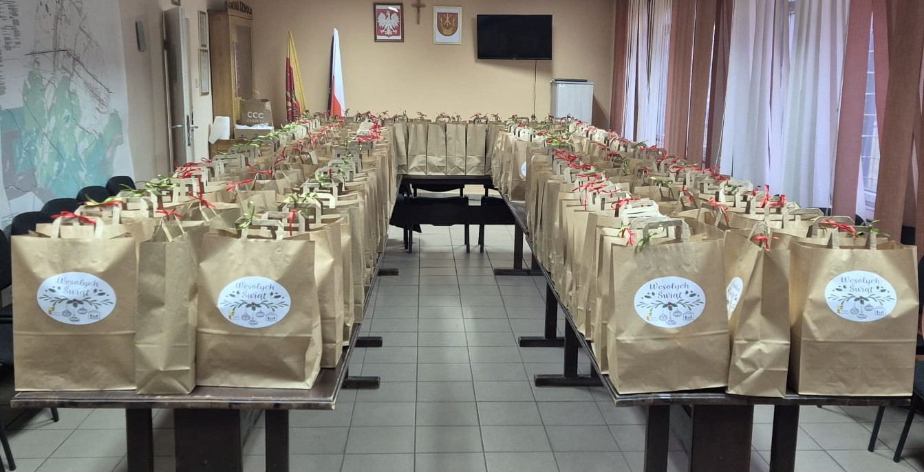 Brązowe papierowe torby z kolorowymi wstążkami i okrągłymi naklejkami z napisem "Wesołych Świąt" ułożone w rzędach na dwóch stołach w jasnym pomieszczeniu z flagami i telewizorem.