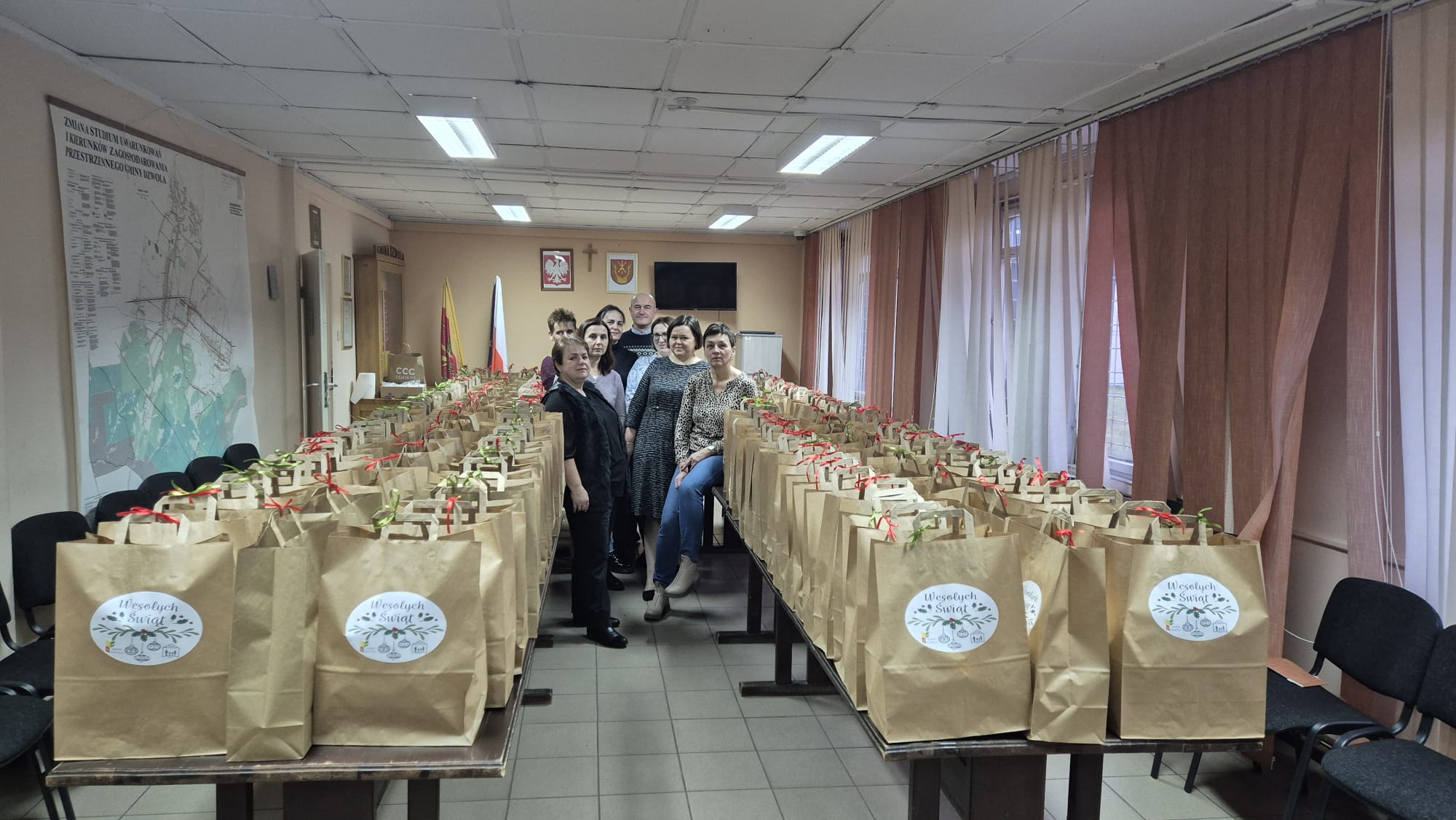 Sześć osób stoi i siedzi między dwoma rzędami stołów z papierowymi torbami ozdobionymi naklejkami „Wesołych Świąt” i czerwonymi oraz zielonymi wstążkami.