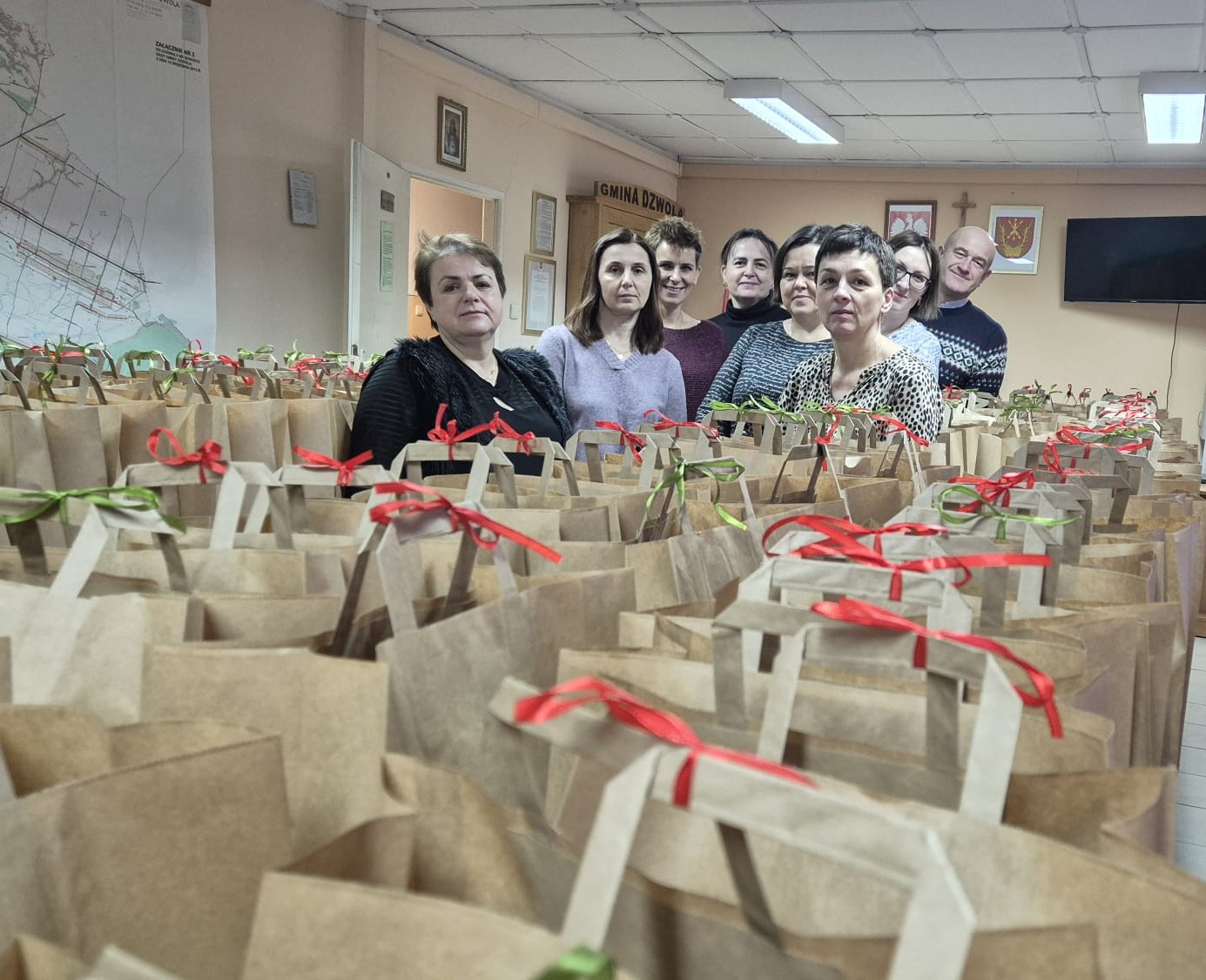Siedem osób za stołem wypełnionym brązowymi papierowymi torbami z czerwonymi i zielonymi wstążkami, jasne pomieszczenie z mapą i telewizorem na ścianie.