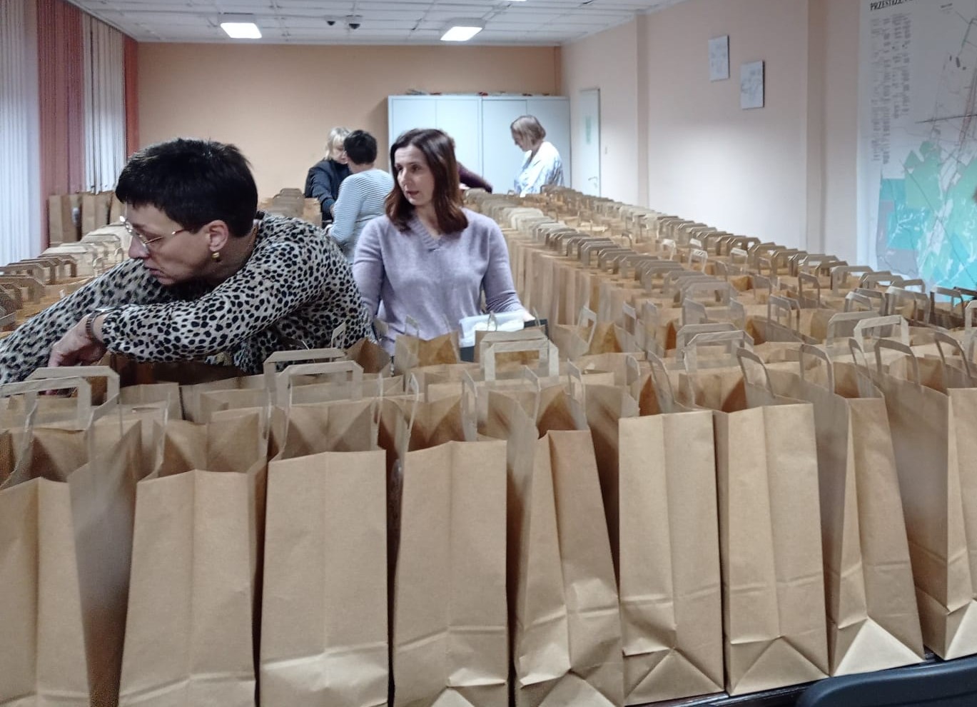 Kobiety stojące przy stołach z wieloma brązowymi papierowymi torbami z uchwytami w jasnym pomieszczeniu biurowym.