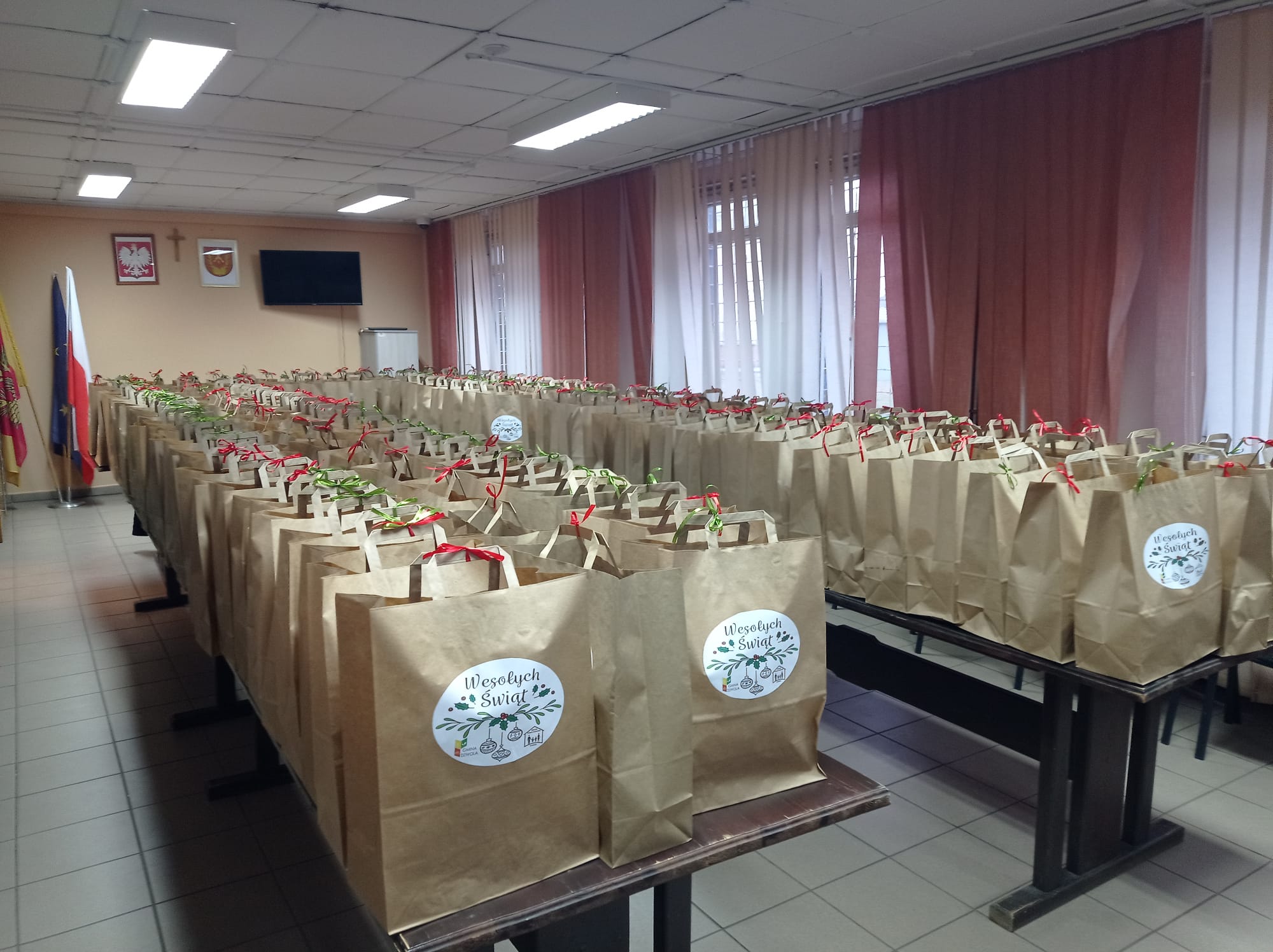 Brązowe papierowe torby z kolorowymi wstążkami i naklejkami "Wesołych Świąt" ułożone w rzędach na stołach w jasnym pomieszczeniu z czerwonymi zasłonami i flagami w rogu.