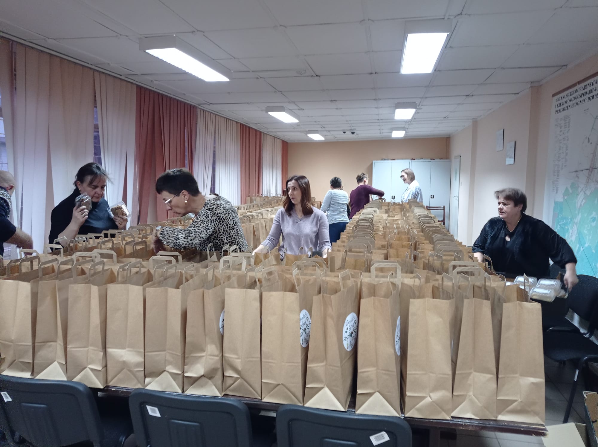 Wnętrze pomieszczenia z kobietami pakującymi produkty do wielu brązowych papierowych toreb z uchwytami, z tyłu jasne zasłony i sufit z kwadratowych paneli.