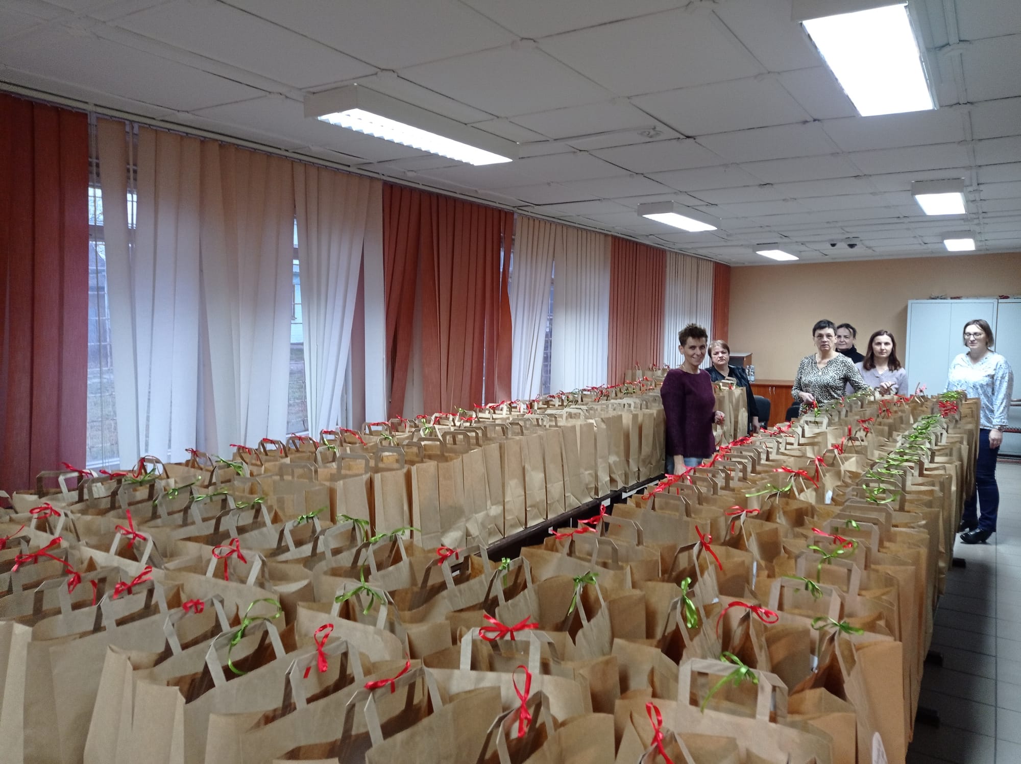 Wnętrze z białym sufitem i oświetleniem, długie stoły z wieloma brązowymi papierowymi torbami z czerwonymi i zielonymi wstążkami, pięć stojących kobiet.