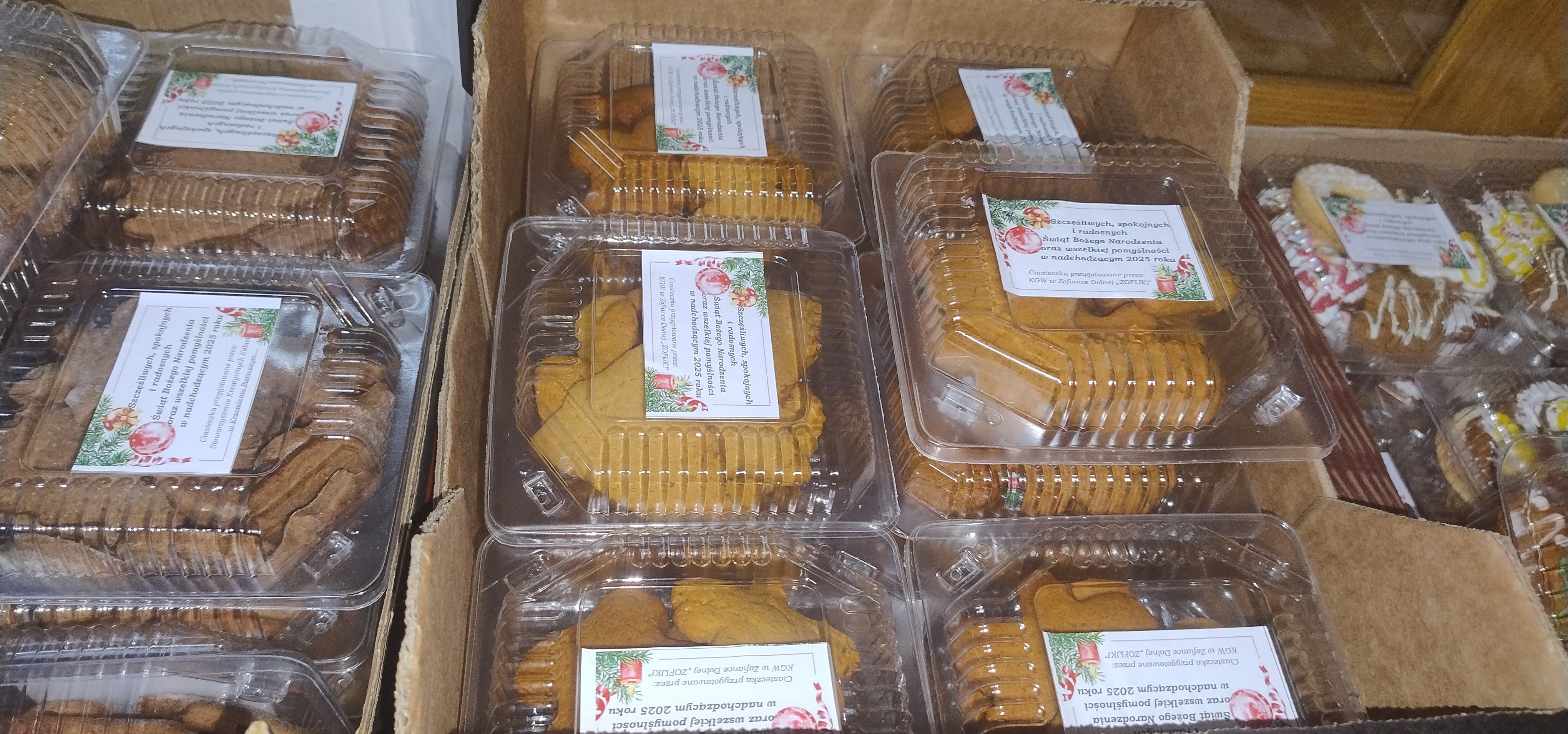 Przezroczyste plastikowe pojemniki z różnymi rodzajami ciastek, niektóre z nich mają kolorowy lukier, wszystkie opakowane i ułożone w kartonowych pudełkach.