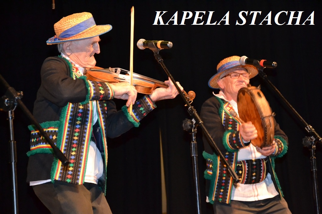 Dwóch starszych mężczyzn w tradycyjnych kolorowych strojach i słomkowych kapeluszach gra na skrzypcach i bębnie, na czarnym tle z białym napisem KAPELA STACHA.
