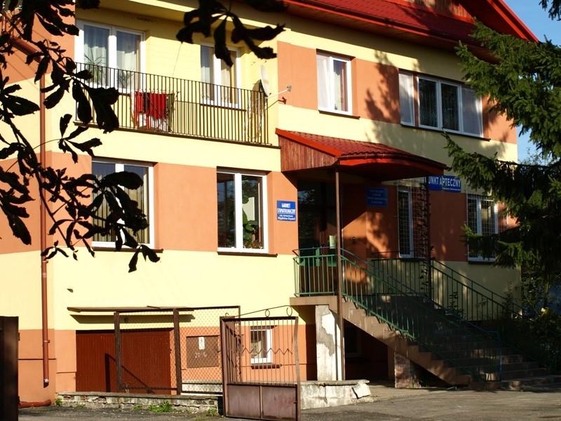 Dwupiętrowy budynek z żółto-brązową elewacją, czerwonym dachem, zielonymi barierkami i schodami, z metalową bramą i drzewami na pierwszym planie.